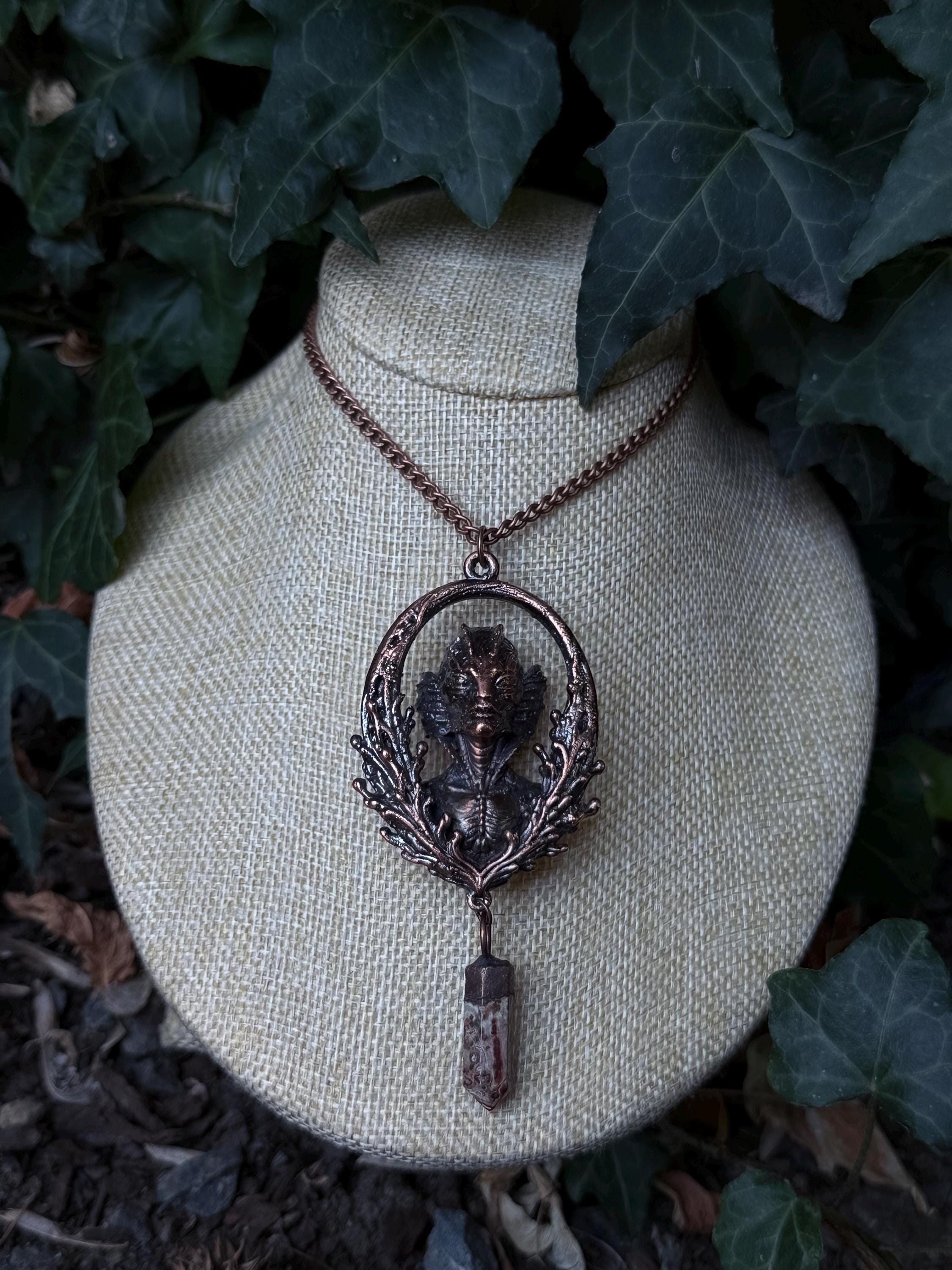 May include: A copper-toned pendant necklace featuring an oval frame with an intricate aquatic creature bust at the center, surrounded by detailed foliage motifs. A small, faceted, brownish-red crystal point hangs from the bottom of the frame.