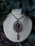 May include: A copper-toned pendant necklace featuring an oval frame with an intricate aquatic creature bust at the center, surrounded by detailed foliage motifs. A small, faceted, brownish-red crystal point hangs from the bottom of the frame.