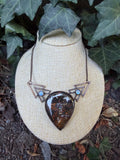 May include: A copper-toned necklace featuring a large, teardrop-shaped pendant with a dark, mottled brown and iridescent surface. The pendant is flanked by two geometric, triangular metal frames, each accented with a small, round, pale blue cabochon.