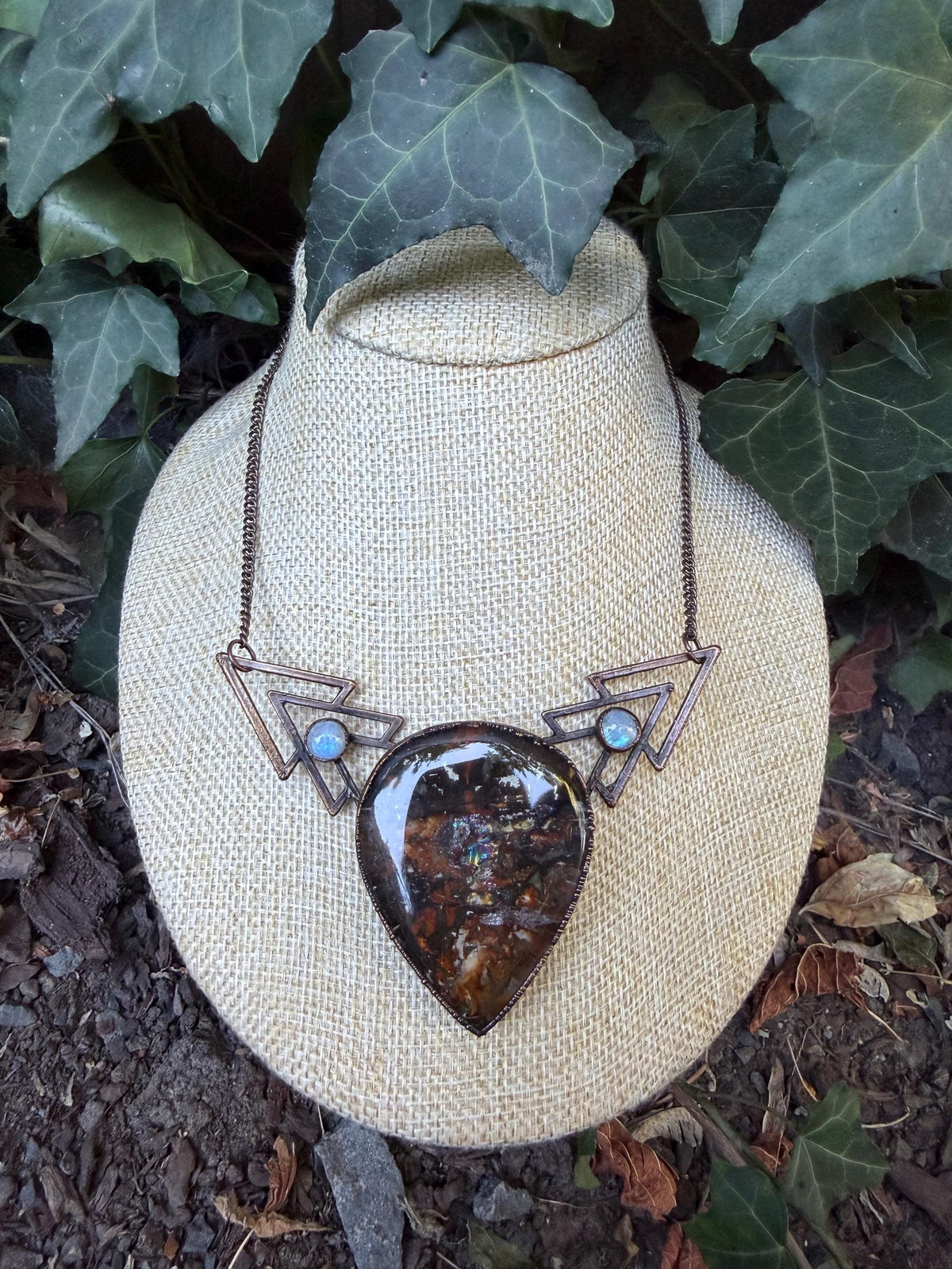 May include: A copper-toned necklace featuring a large, teardrop-shaped pendant with a dark, mottled brown and iridescent surface. The pendant is flanked by two geometric, triangular metal frames, each accented with a small, round, pale blue cabochon.