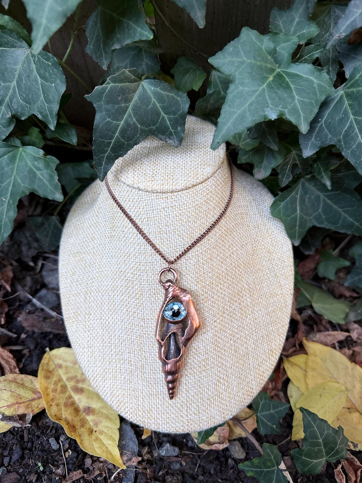 May include: A copper-toned pendant necklace featuring a spiral shell casing with a metallic finish. The pendant is centered with a single, round, light blue glass eye cabochon. The piece hangs from a matching copper-colored metal chain.